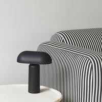 Black mushroom lamp on a white table beside a black and white striped sofa in a minimalist setting.