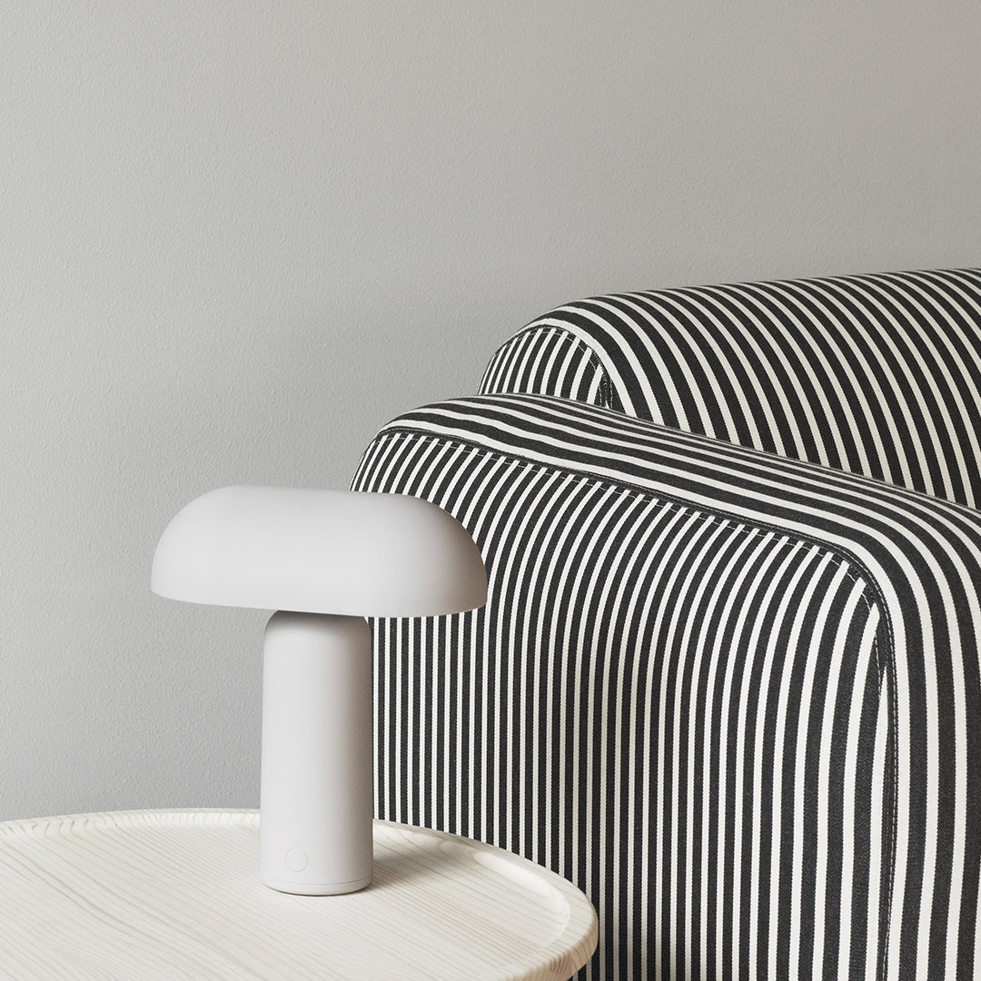 Gray backdrop with a black and white striped sofa and a minimalist white lamp on a light wood table.