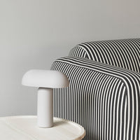 Gray backdrop with a black and white striped sofa and a minimalist white lamp on a light wood table.