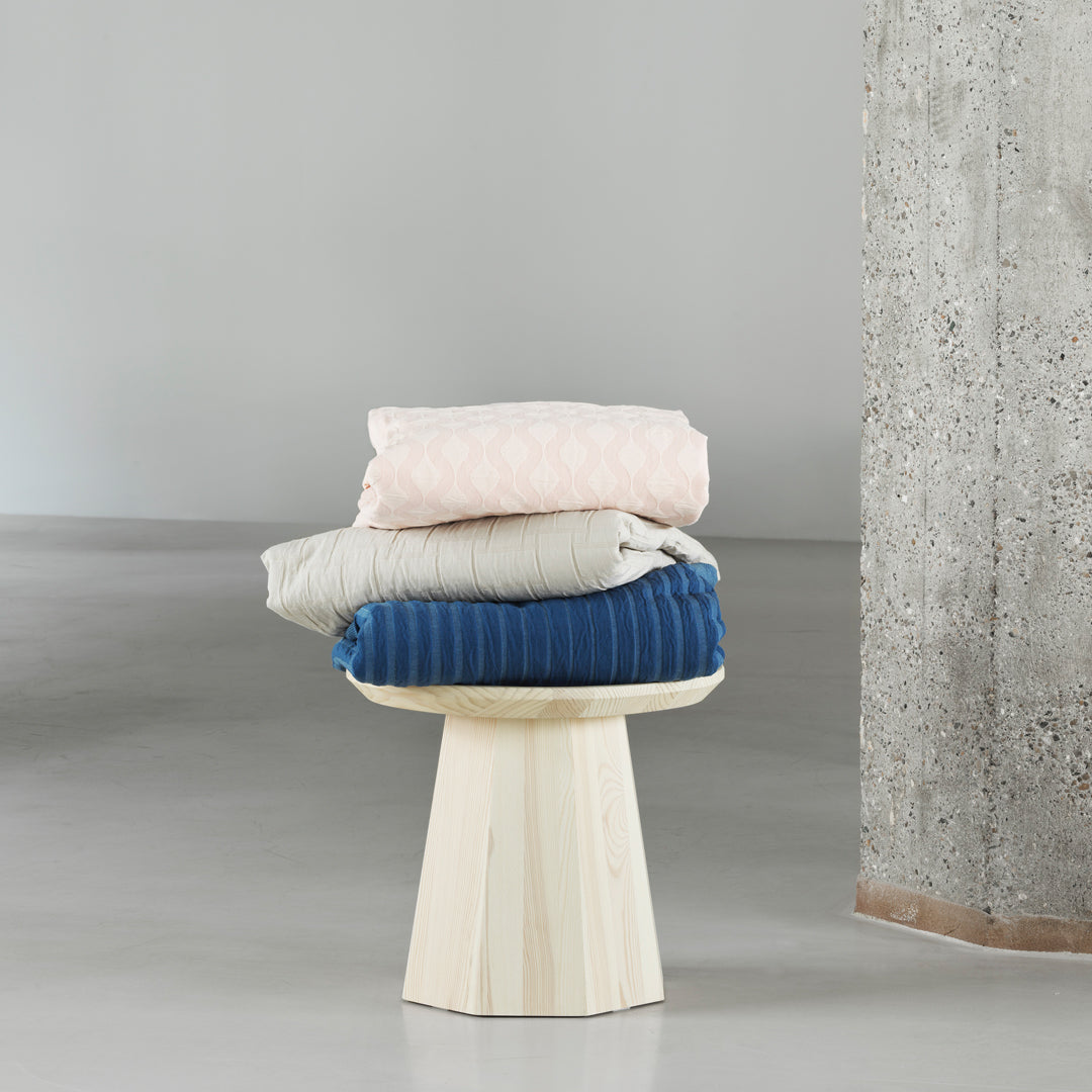 Soft pink, cream, and deep blue bedding with subtle patterns, neatly stacked on a natural wood stool.