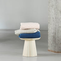 Soft pink, cream, and deep blue bedding with subtle patterns, neatly stacked on a natural wood stool.