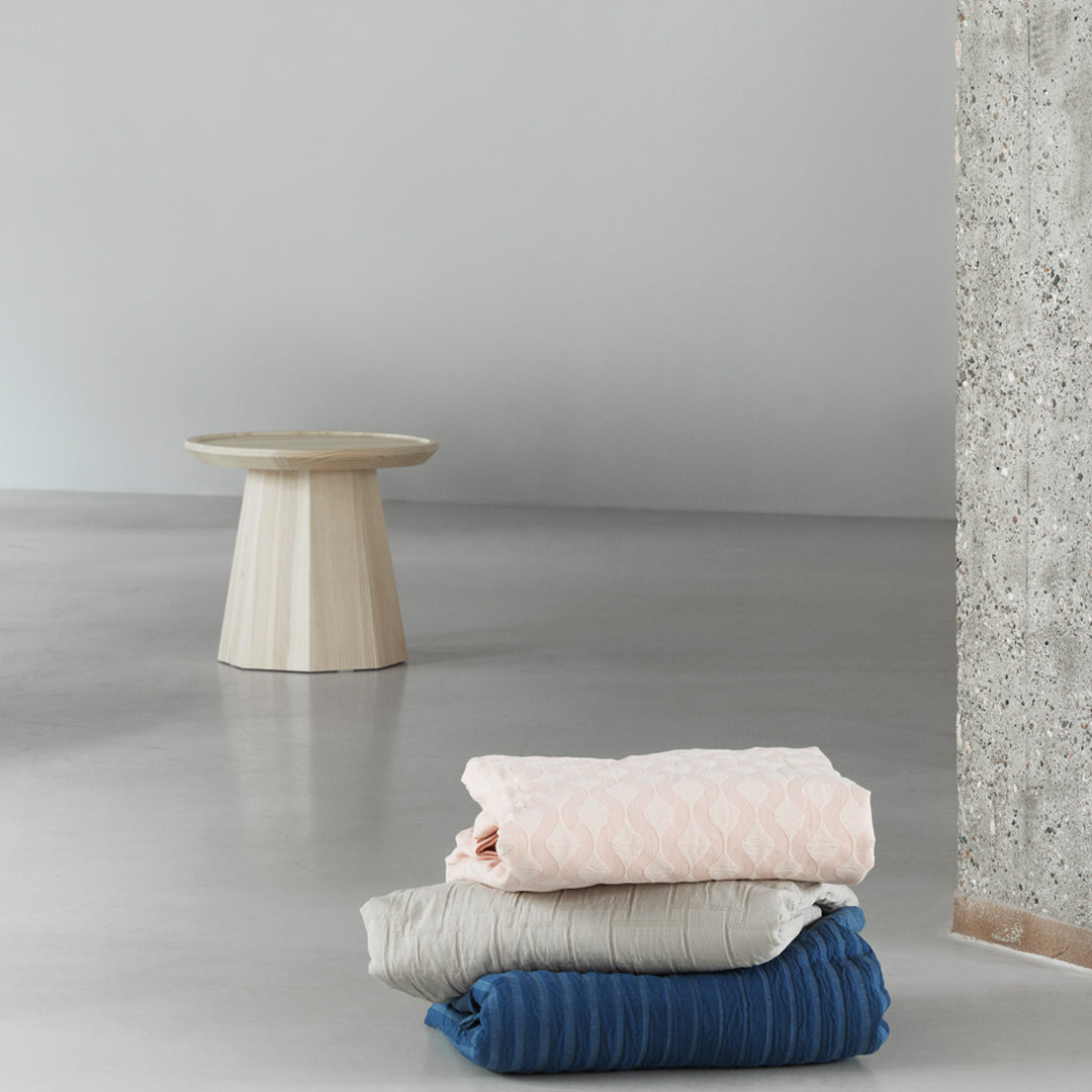 Stacked blankets in pastel pink, soft gray, and deep blue with textured patterns, against a minimalistic gray backdrop.