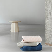 Stacked blankets in pastel pink, soft gray, and deep blue with textured patterns, against a minimalistic gray backdrop.