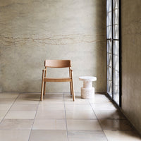 "Minimalist wooden chair with a light tan cushion against a textured grey wall and smooth beige tiles."