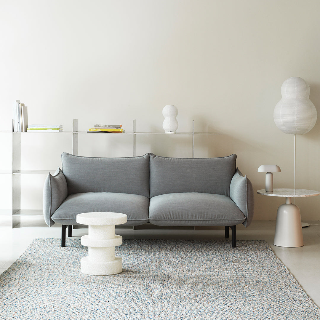 Light gray sofa with simple stitching, set against a pale wall and patterned blue area rug. Modern decor accents in soft hues.