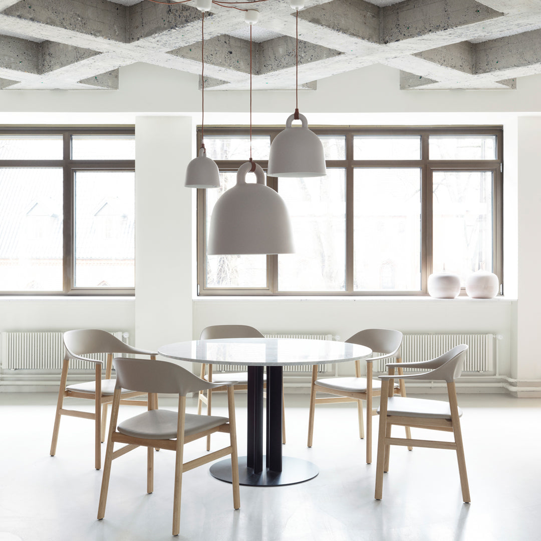 Modern dining room with a round white table, natural wood chairs, and gray pendant lights against a clean, minimal backdrop.