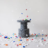 Gray speckled pedestal with colorful scattered confetti in red, blue, yellow, and white on a white background.
