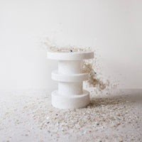 White three-tiered pedestal with a textured surface, surrounded by multicolored glitter particles.
