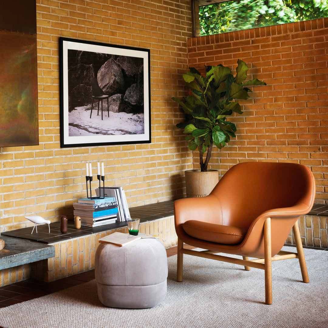 "Modern brown leather chair and soft gray pouf in a warm brick setting, complemented by greenery and artistic decor."