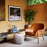 "Modern brown leather chair and soft gray pouf in a warm brick setting, complemented by greenery and artistic decor."