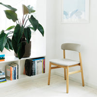 Light gray upholstered chair with natural wood legs, set against a bright, airy room with greenery and books.