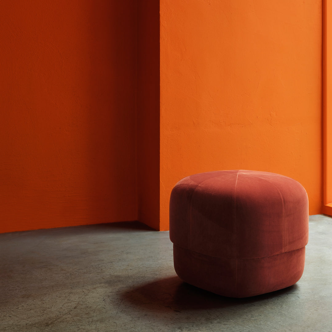 Soft velvet pouf in a rich terracotta color, set against a vibrant orange wall.