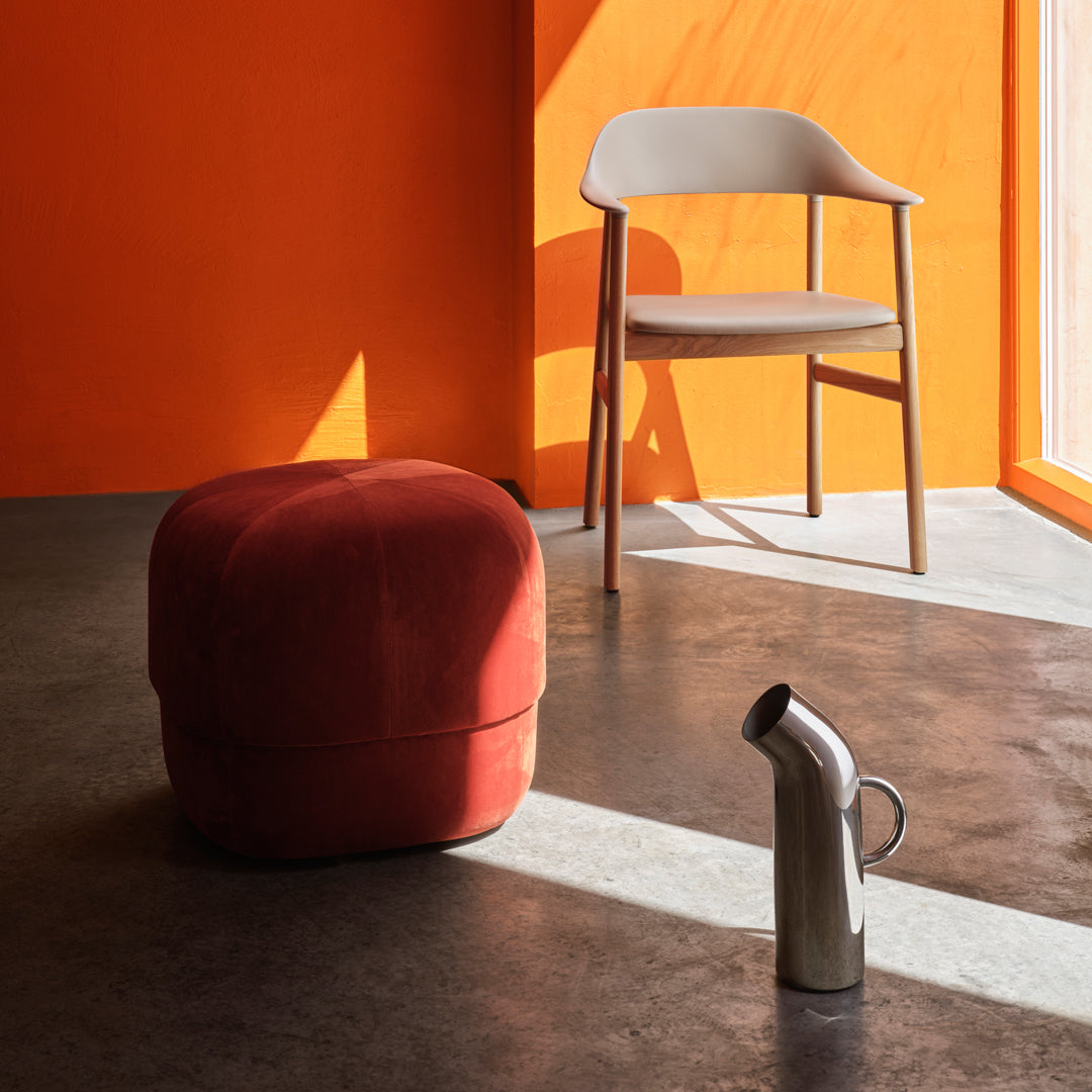 Red velvet pouf with orange wall, gray chair, and silver pitcher in a sunny, modern interior.