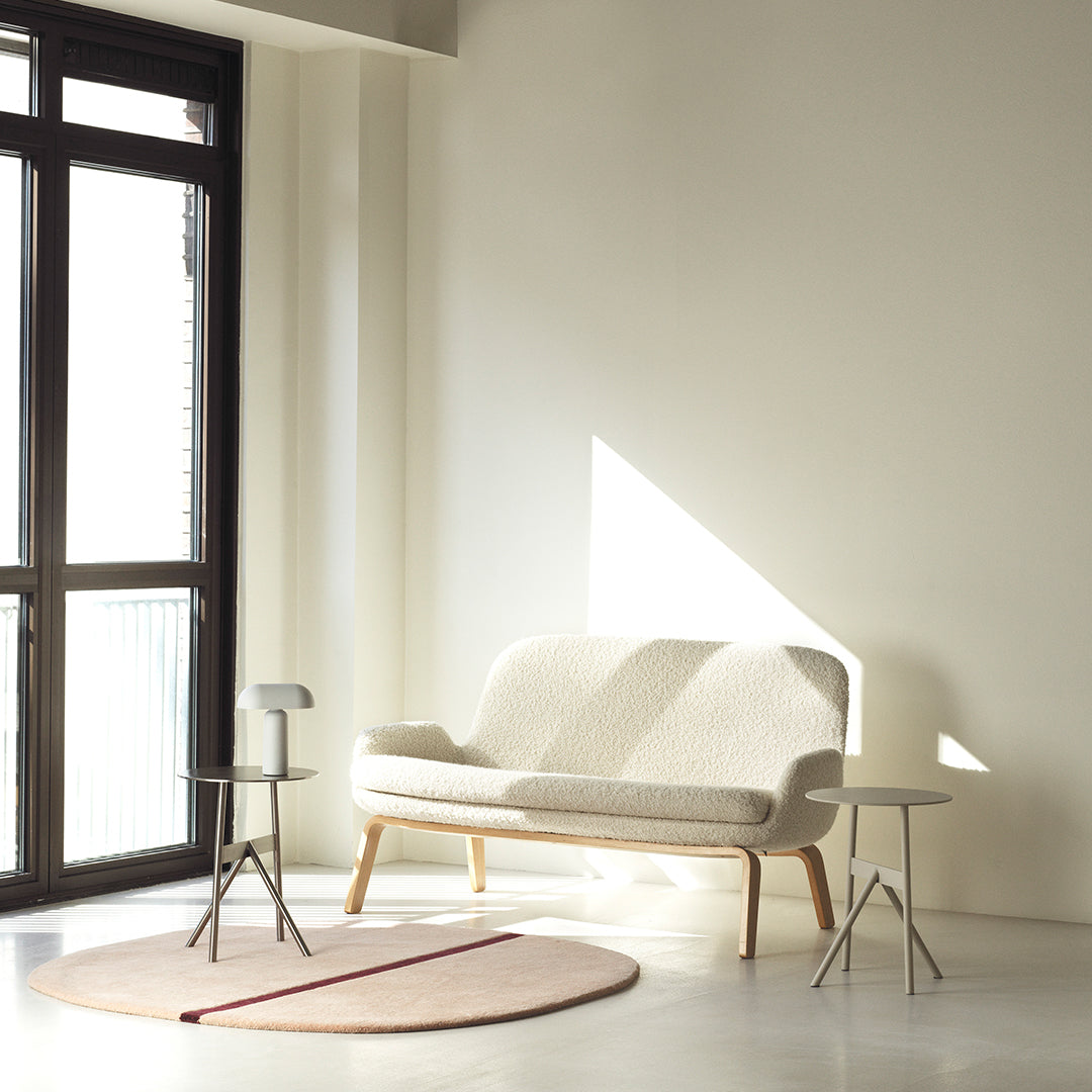 Light beige fabric sofa on wooden legs with a round pale pink rug and minimalist side tables, set in a bright room.