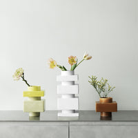 Stacked geometric vases in white, green, and terracotta with floral arrangements, showcasing modern design and soft colors.