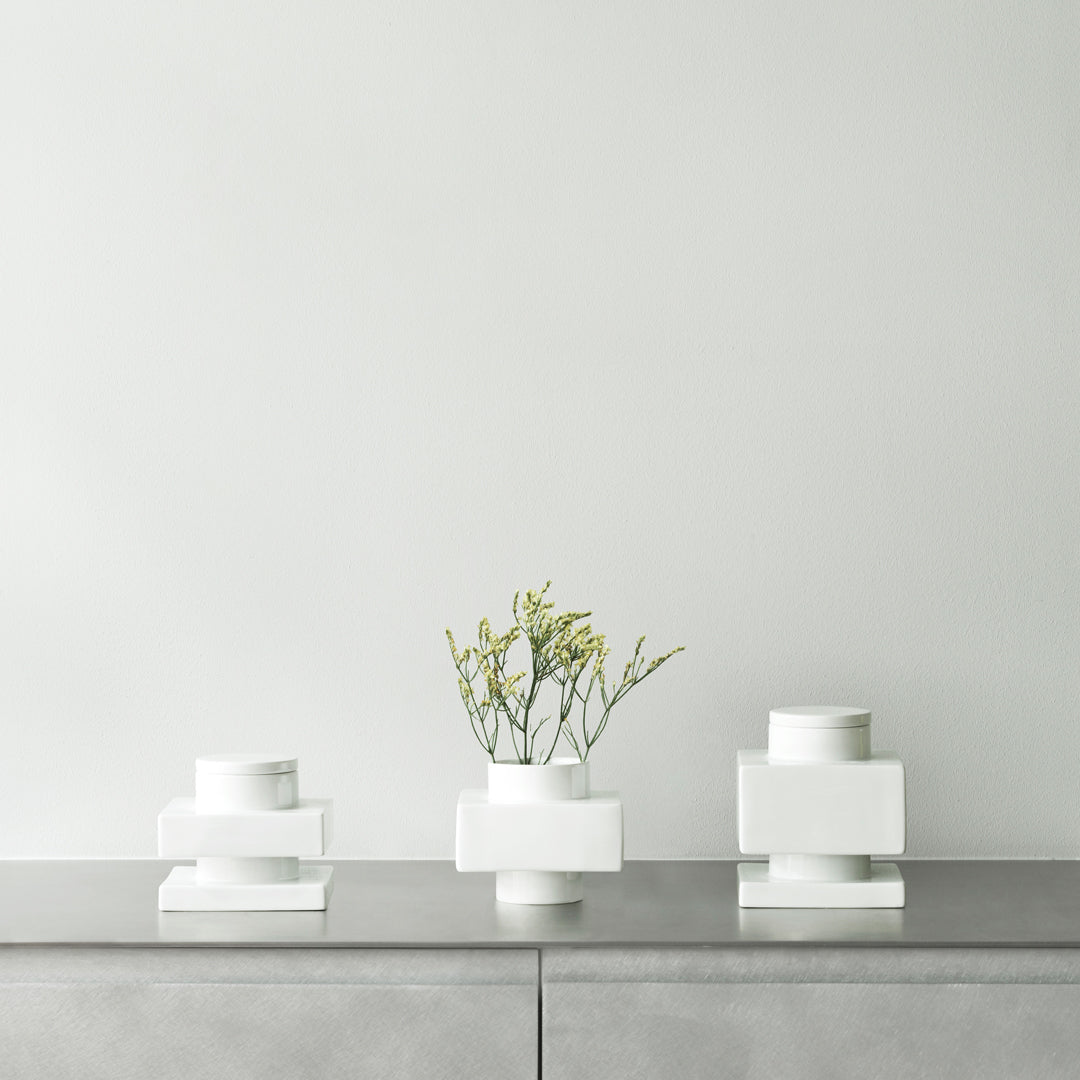 Three minimalist white vases with geometric shapes on a sleek gray surface, featuring subtle greenery.