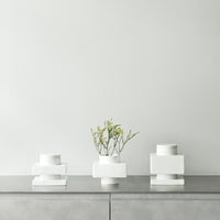 Three minimalist white vases with geometric shapes on a sleek gray surface, featuring subtle greenery.