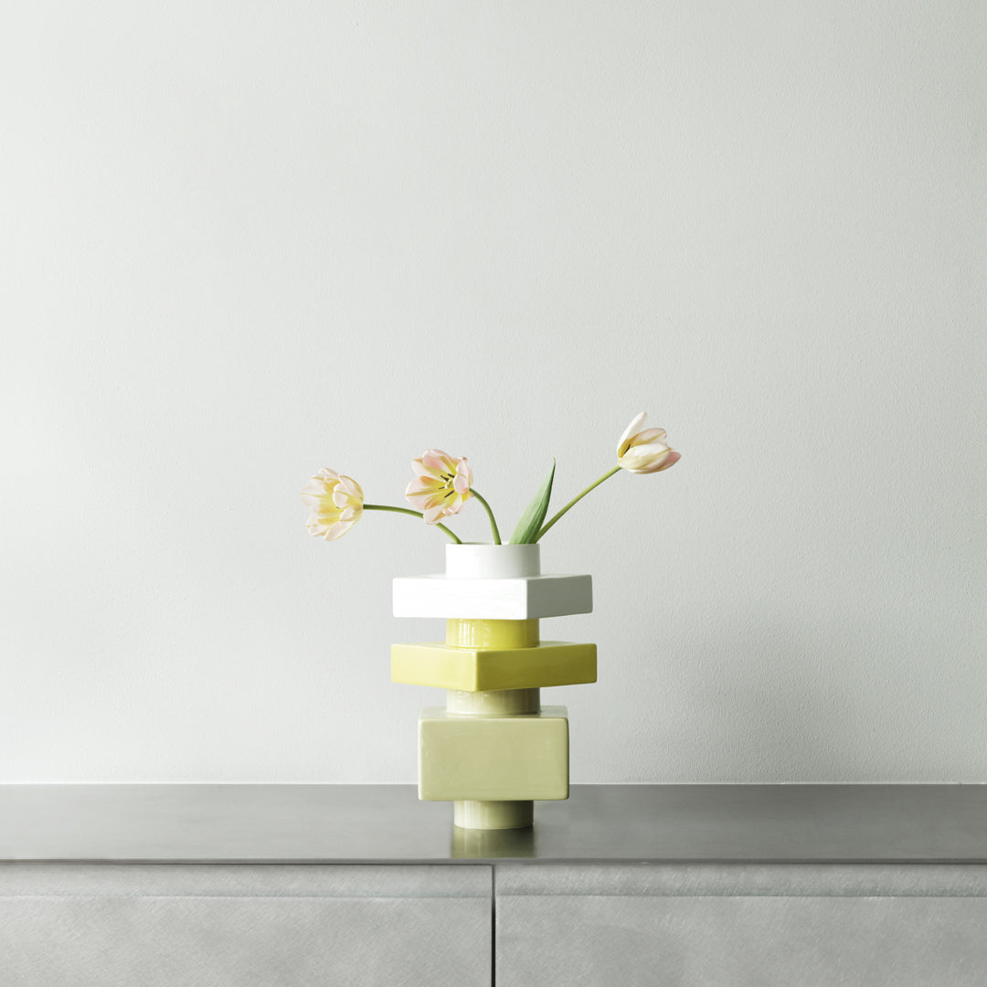 Abstract stacked vase in white, yellow, and olive colors, adorned with delicate pink flowers against a neutral background.