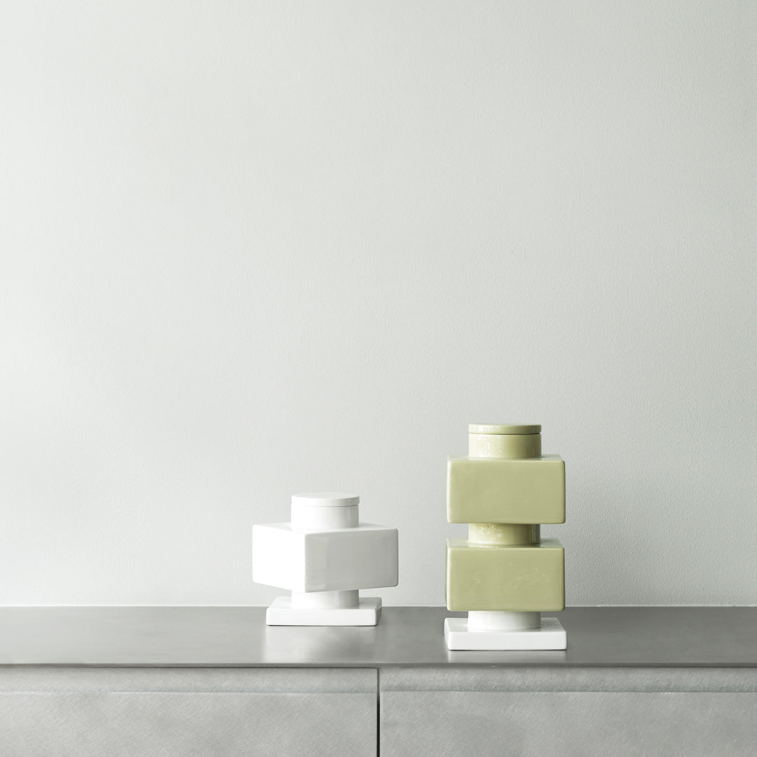 Stacked geometric vases in white and soft green on a minimalist gray surface against a neutral backdrop.