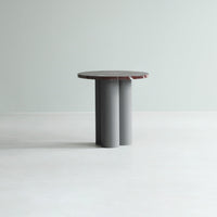 Round table with a gray base and a textured, dark red top against a light green background. Minimalist design.