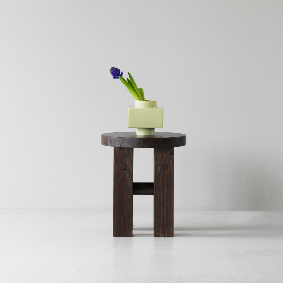 Dark wood table with a round top, featuring a pale yellow geometric vase and green foliage, set against a light backdrop.