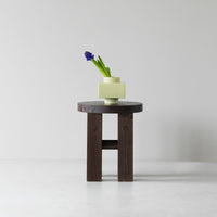 Dark wood table with a round top, featuring a pale yellow geometric vase and green foliage, set against a light backdrop.