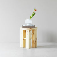 Light wood stool with smooth surface and subtle grain, topped with a white geometric object and green flower.