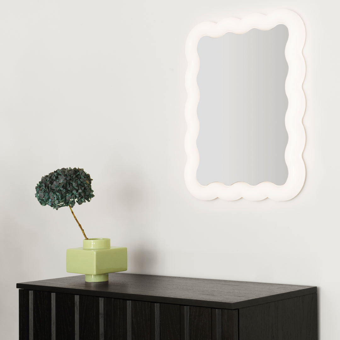 "Soft white scalloped mirror on a light wall, paired with a green planter on a black cabinet."