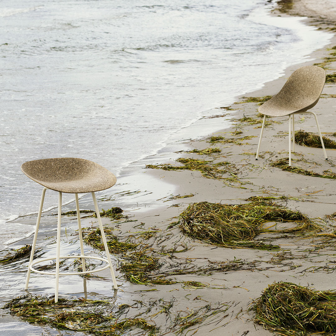 Neutral beige bar stools with a smooth, textured surface, set against a coastal backdrop of sand and sea.