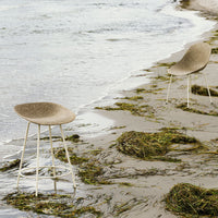Neutral beige bar stools with a smooth, textured surface, set against a coastal backdrop of sand and sea.