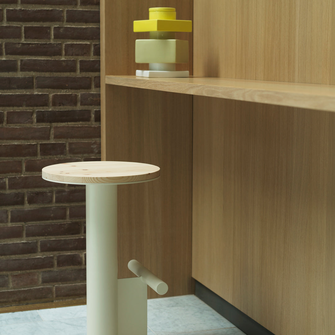 Minimalist light cream stool with a circular wooden top, set against a warm wooden backdrop and brick wall.