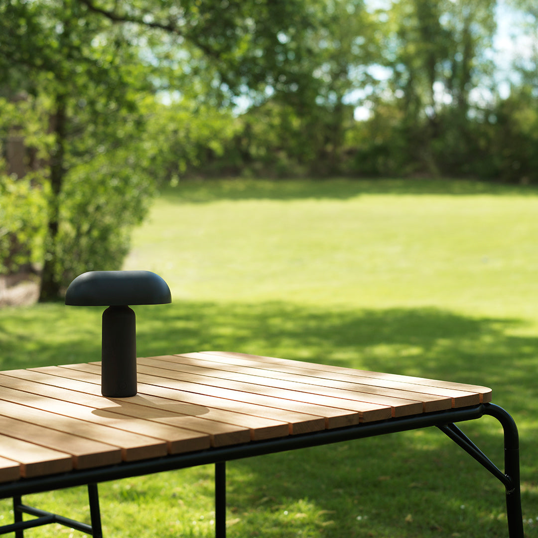 Black table lamp on natural wood surface, set against a vibrant green lawn and dappled sunlight.