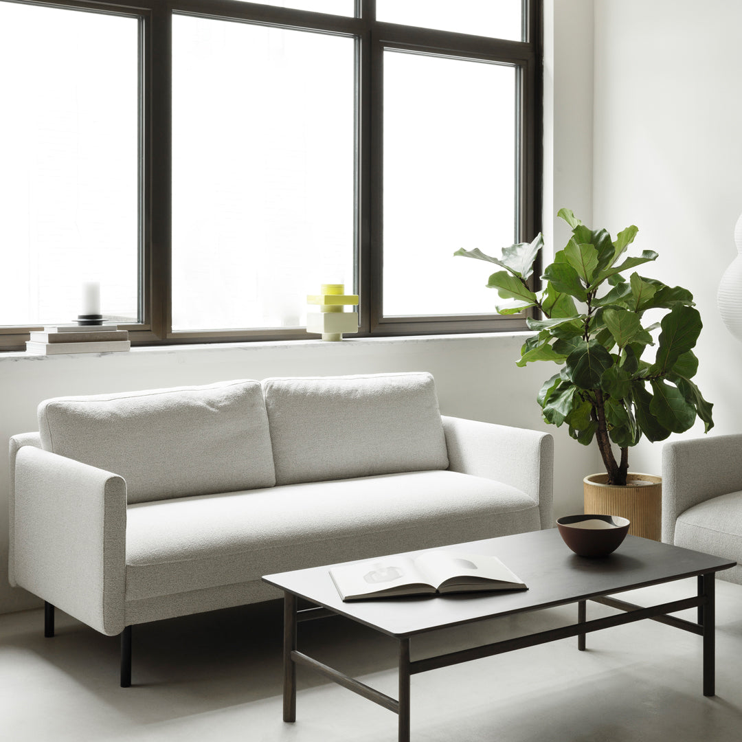Modern white sofa with sleek lines, paired with a dark wooden coffee table, surrounded by greenery. Bright and airy setting.