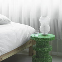 Green speckled side table with a white paper lamp, arranged beside a white bed and soft sheer curtains.