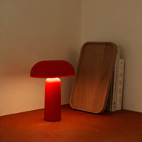 Red mushroom-shaped lamp with a smooth finish, casting a warm glow on a brown surface. Minimalist wooden tray in background.