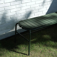 Green padded bench with vertical stripes, set against a white brick wall and grass background.