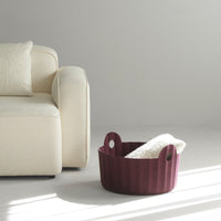 Plum round basket with textured pattern, featuring two handles and a soft white towel inside, near a cream sofa.
