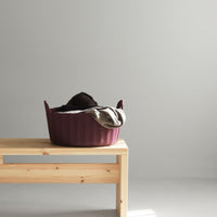 "Plum pet bed with ribbed texture, featuring a cozy gray blanket draped on top, placed on a wooden bench."