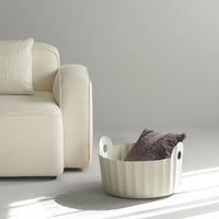 Cream sofa with a textured white cushion next to a pale gray basket holding a deep brown pillow on a light floor.