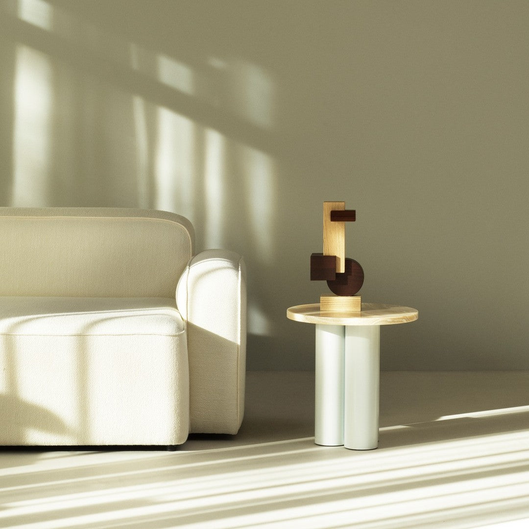 Beige sofa with soft texture, circular wood and white table with geometric wooden sculpture, neutral color palette.
