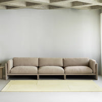 Beige modern three-seater sofa on a light yellow rug, set against a minimalist gray wall and concrete ceiling.