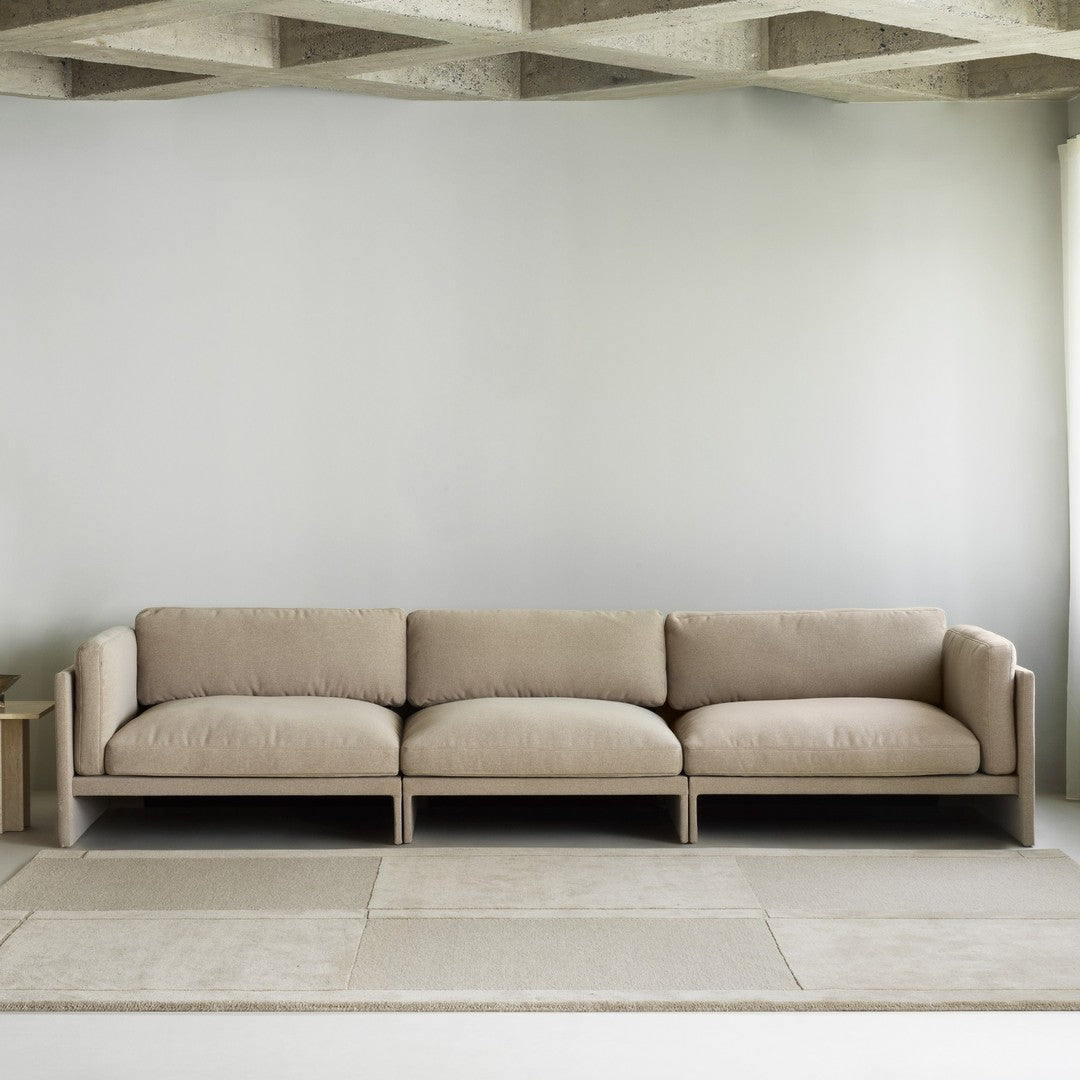 Beige modular sofa with a smooth texture and minimalist design, complemented by a light gray wall and soft rug tones.