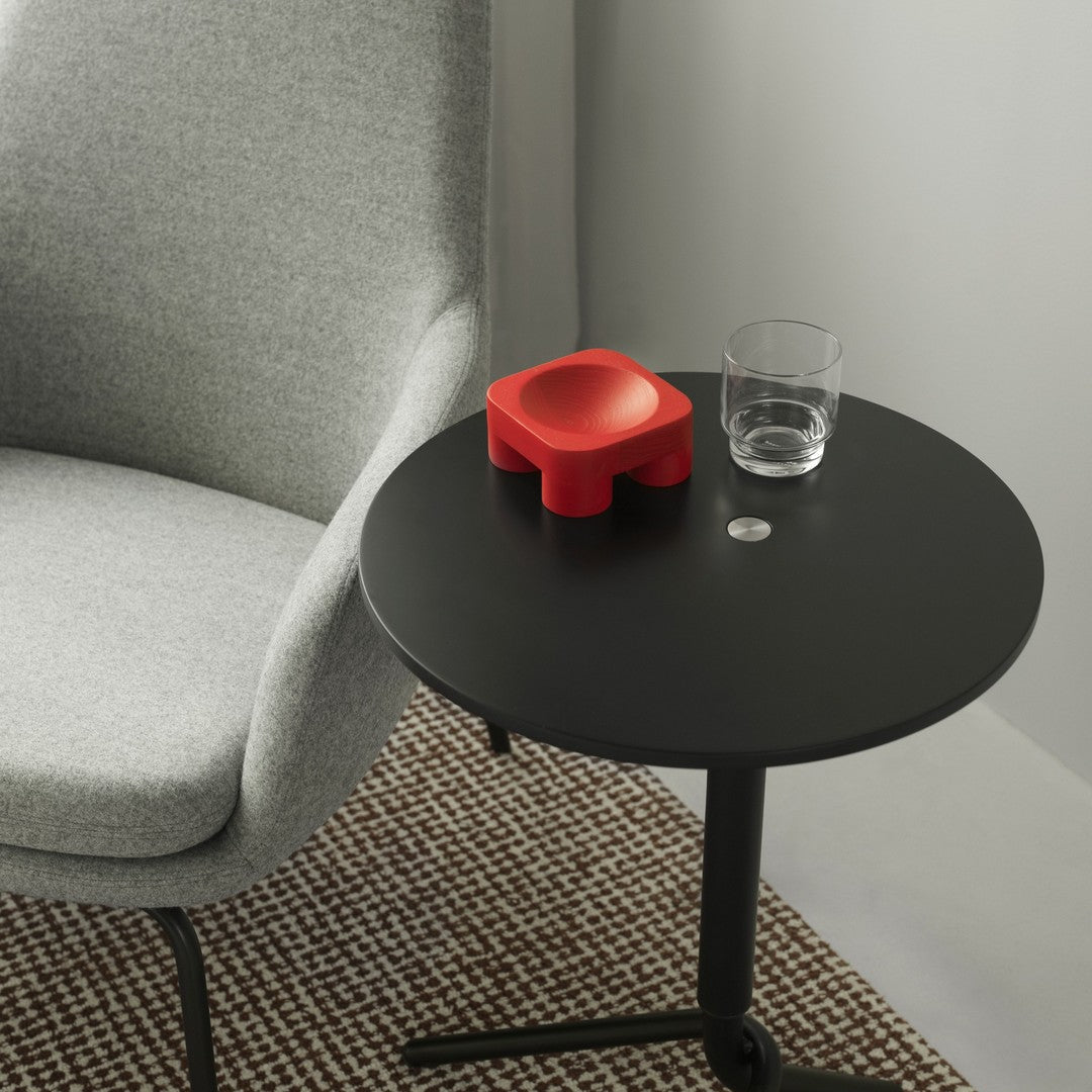 Gray upholstered chair beside a black round table with a bright red object, clear glass, and patterned rug.