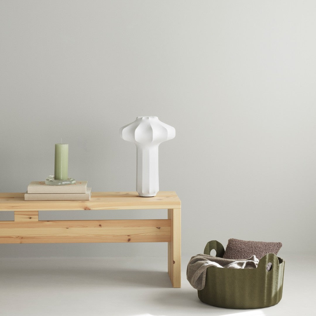 White lamp on a wooden table, green candle, and olive basket with gray blanket; minimalist neutral color palette.