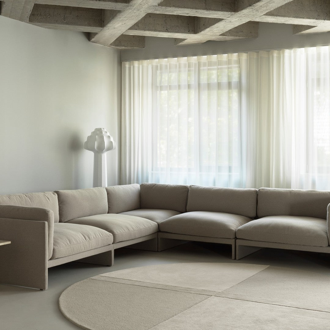 Beige sectional sofa on soft, light gray floor, complemented by sheer white curtains and a minimalist white lamp.