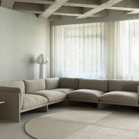Beige sectional sofa on soft, light gray floor, complemented by sheer white curtains and a minimalist white lamp.