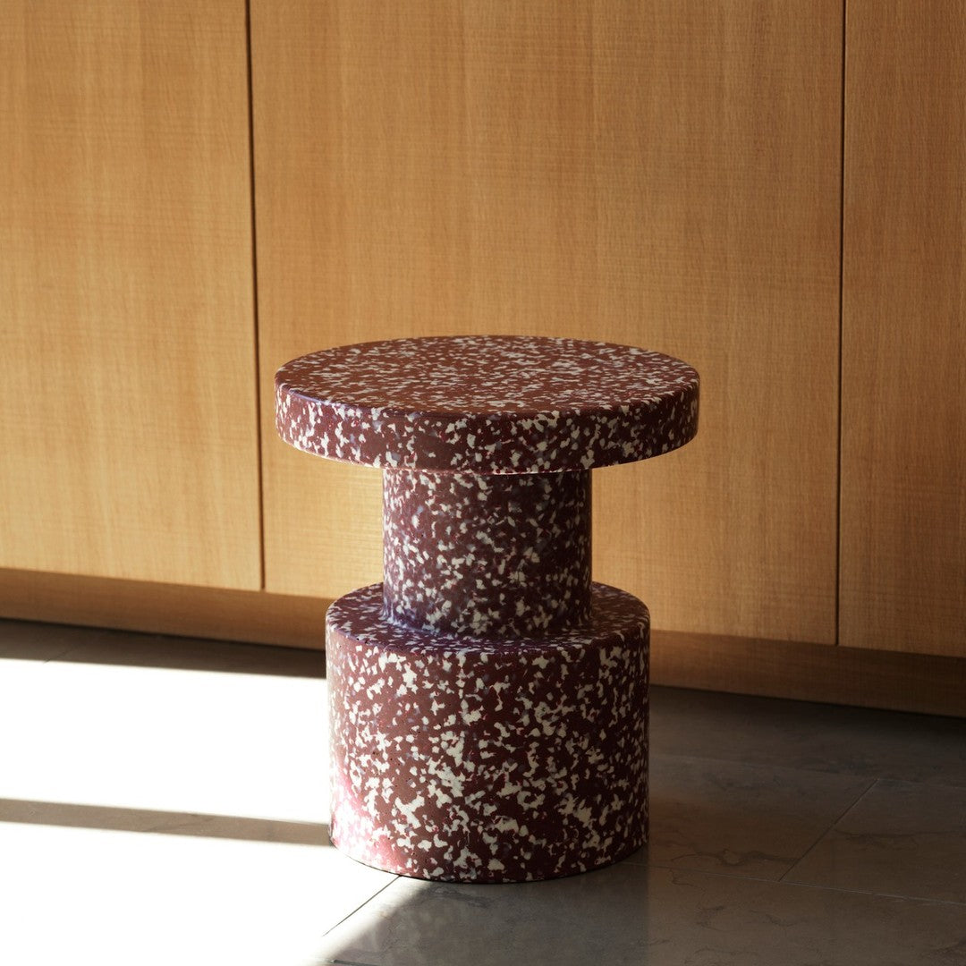 Round maroon table with a speckled white pattern, featuring a cylindrical base and minimalist design.