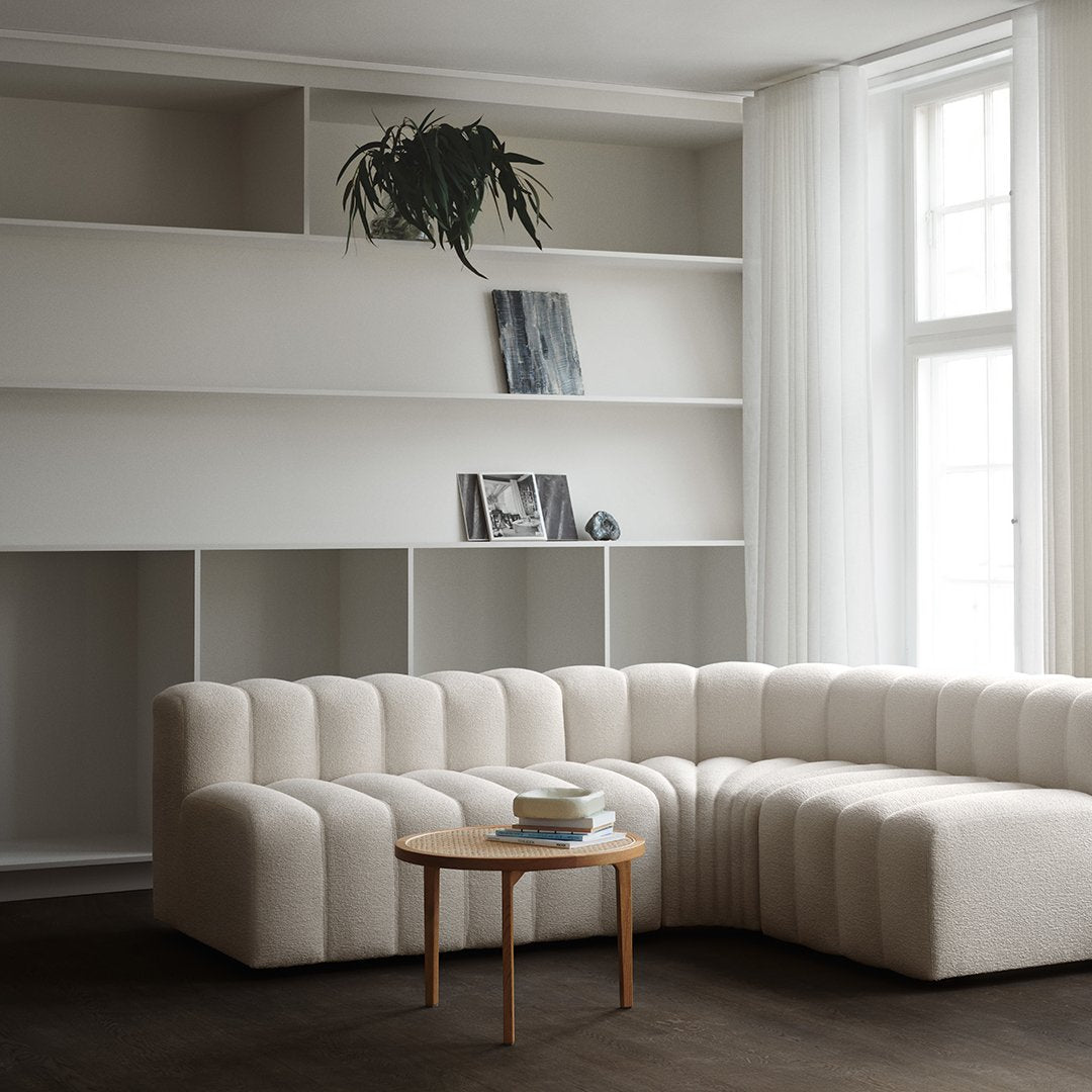 Soft beige curved sofa with plush ribbed texture, set against a minimalist white bookshelf and dark brown floor.