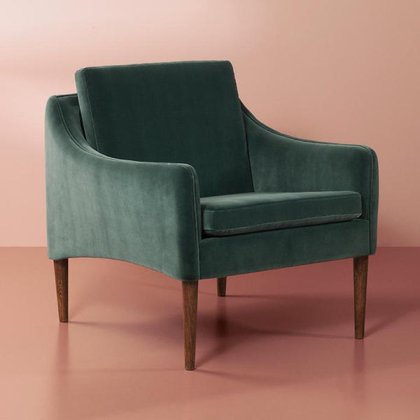 Velvet green armchair with subtle curved design and wooden legs, set against a soft pink background.