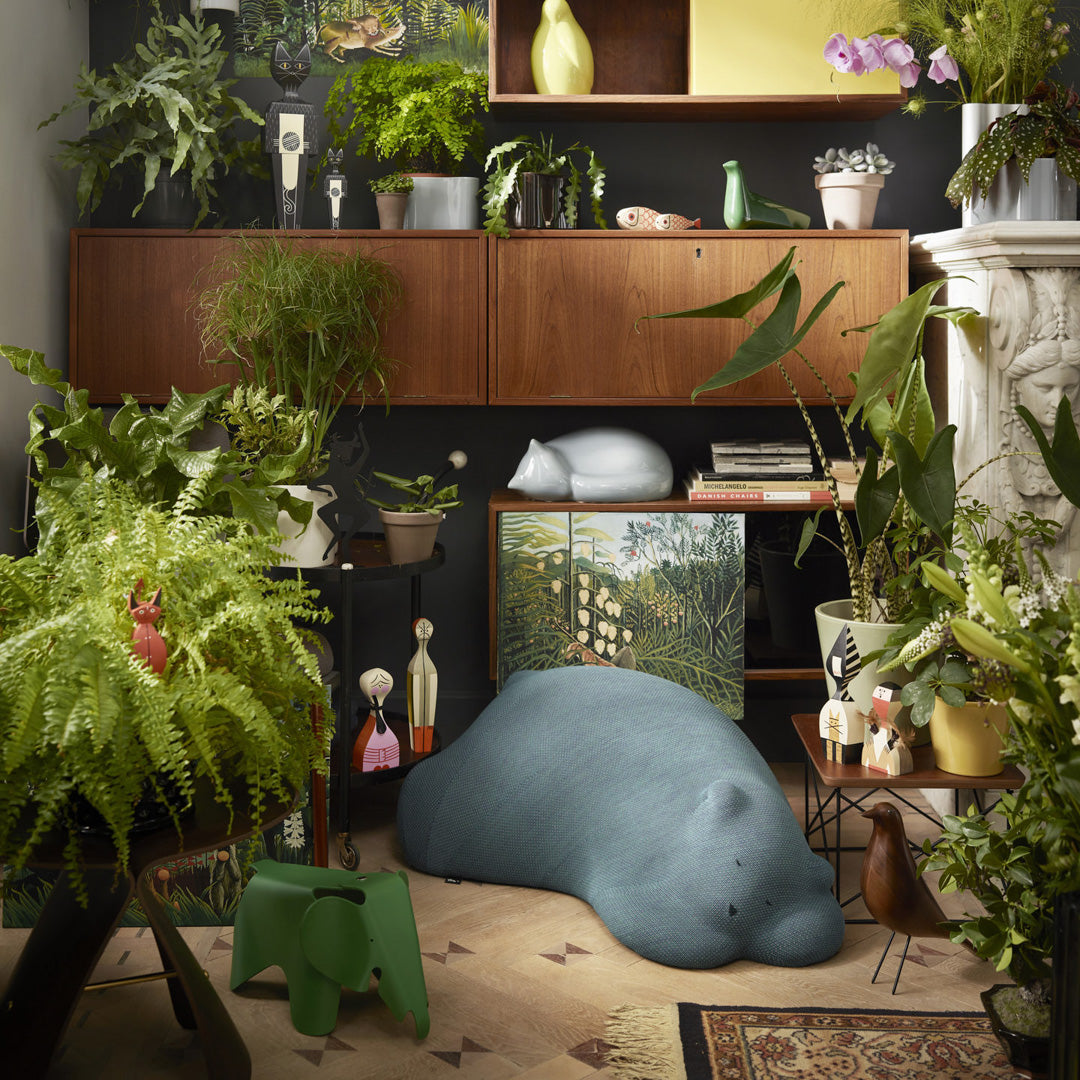 Blue and green cushion shaped like a manatee, surrounded by lush greenery and patterned decor in a cozy interior setting.